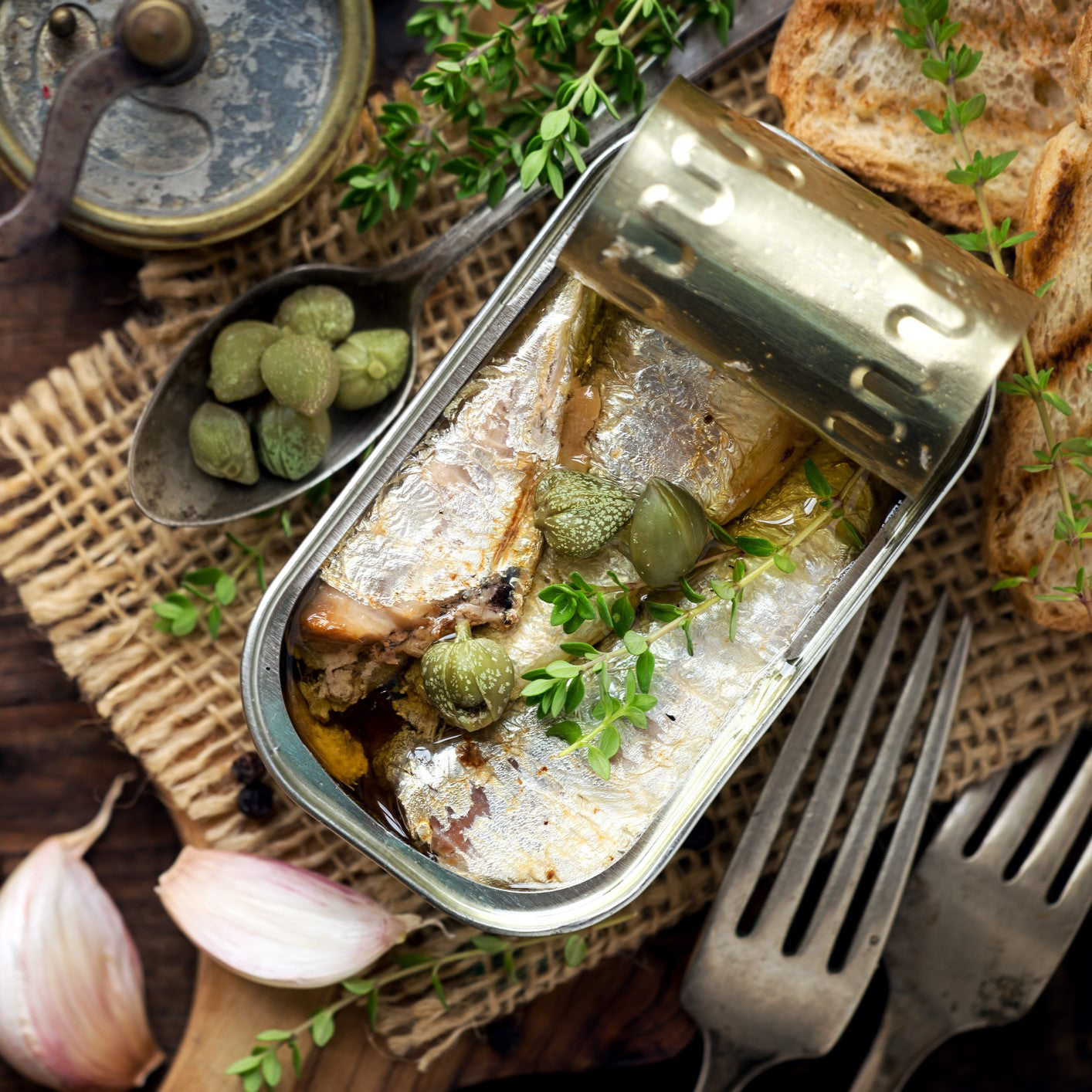 Open tin of sardines in olive oil served with capers, garlic, fresh thyme and toasted bread